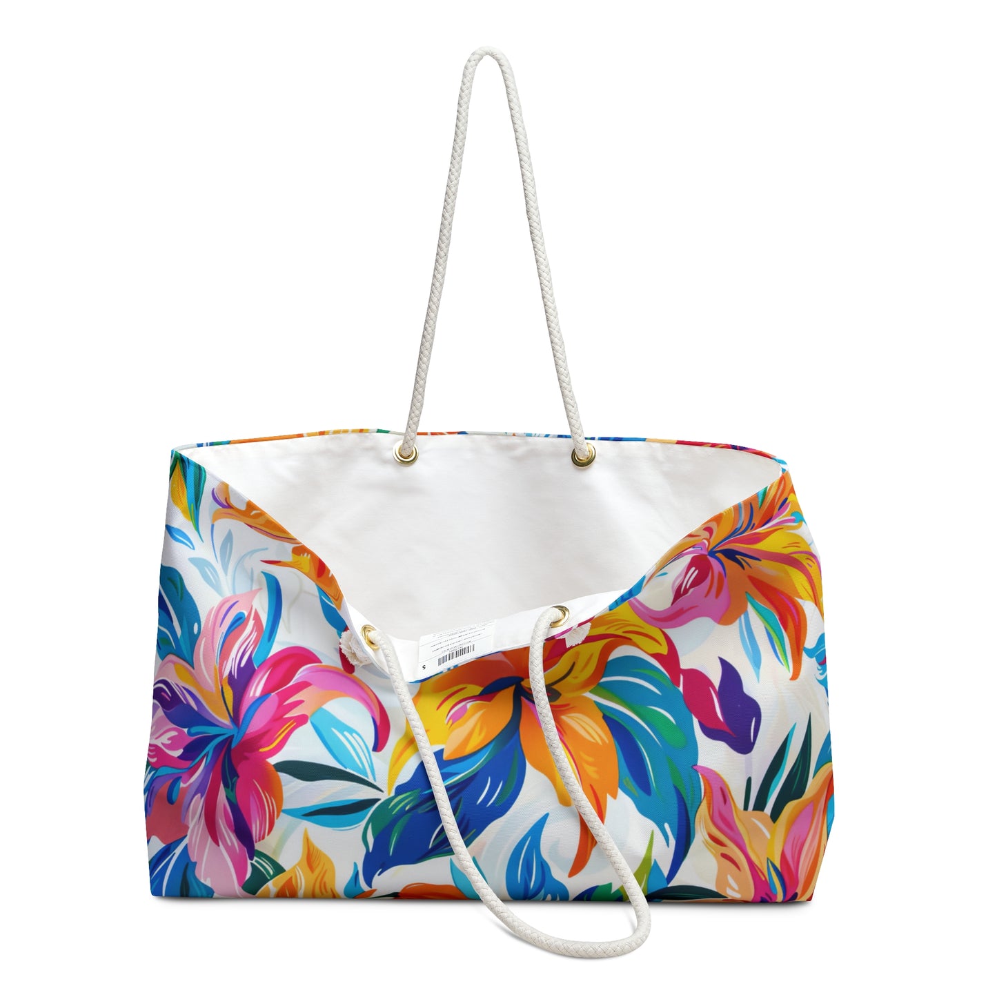 Oversized floral weekender tote bag with colorful tropical flowers, white rope handles, displayed on a sandy beach background opened to display white linen interior