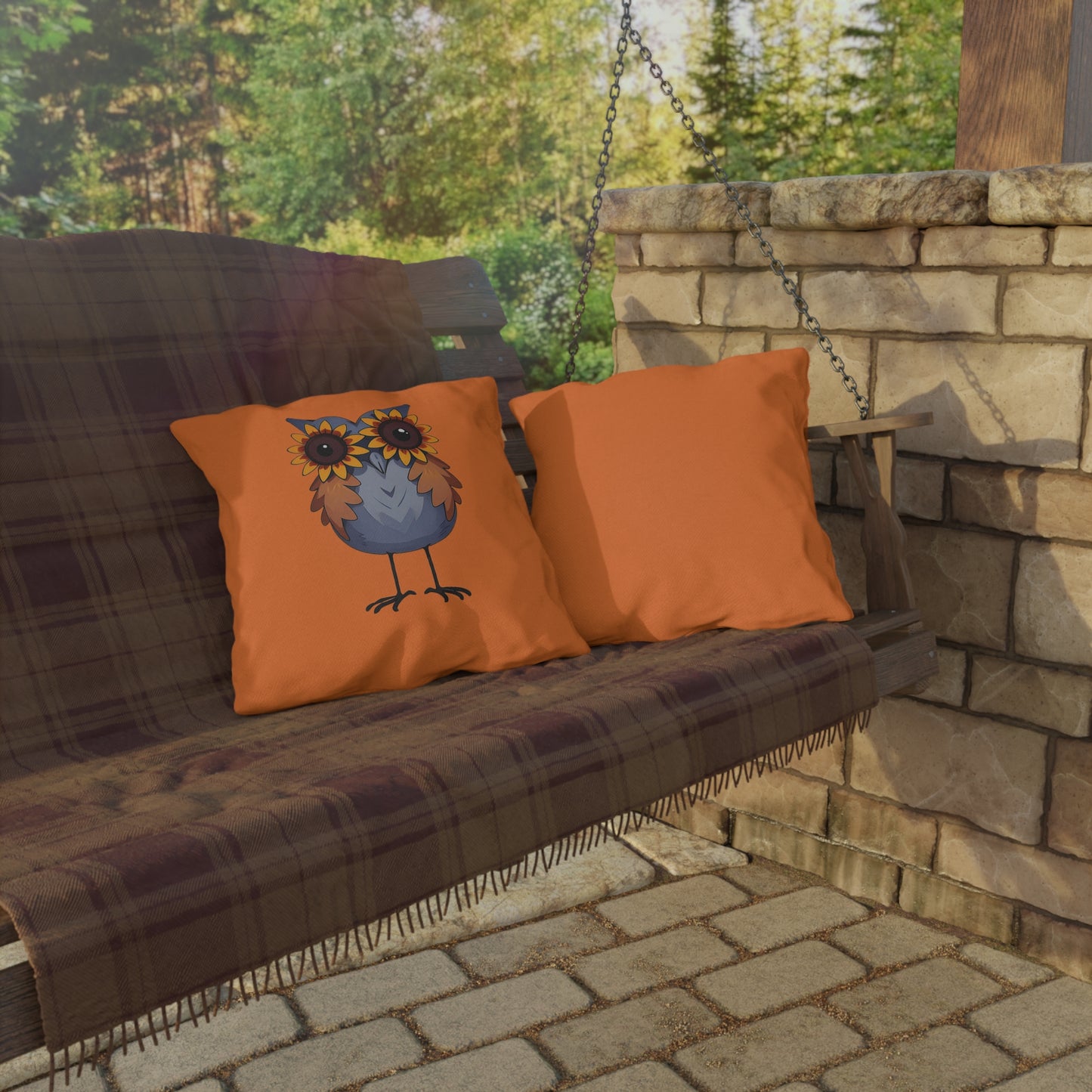 Wooden swing with plaid cover and orange pillows featuring an owl design, set against a stone wall and greenery.