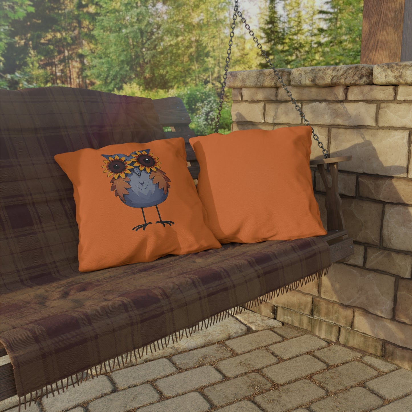 Outdoor swing with plaid seat and orange pillows featuring an owl design, set against a stone wall and greenery.