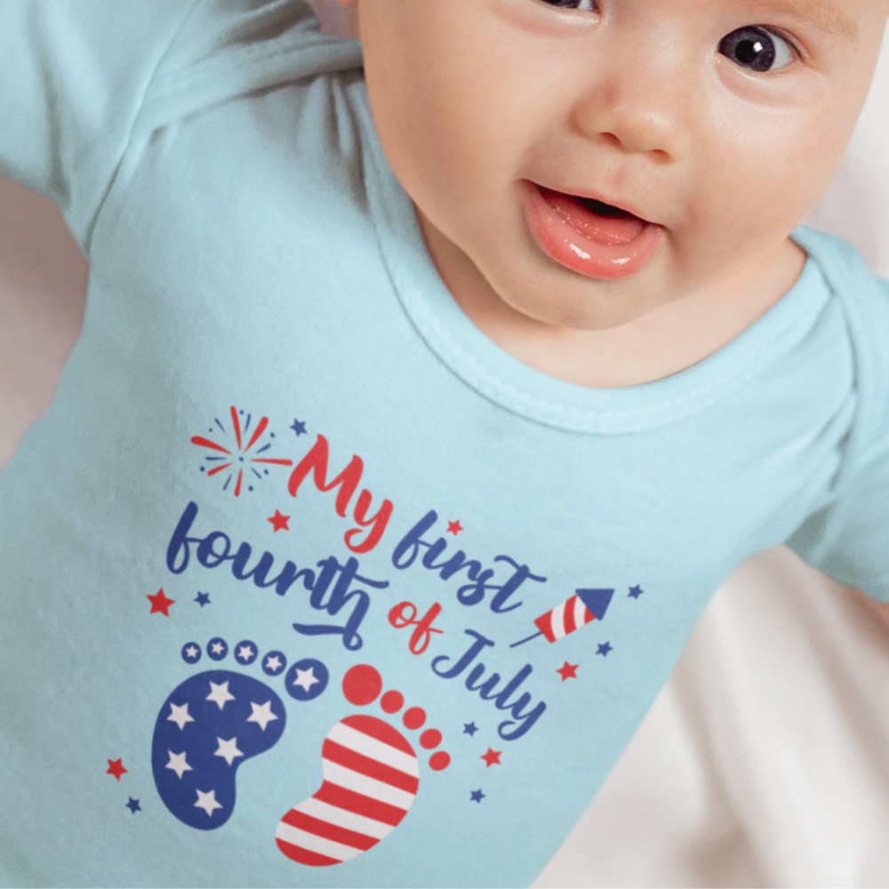 Smiling baby wearing a light blue bodysuit with “My First Fourth of July” design featuring patriotic baby footprints and fireworks.