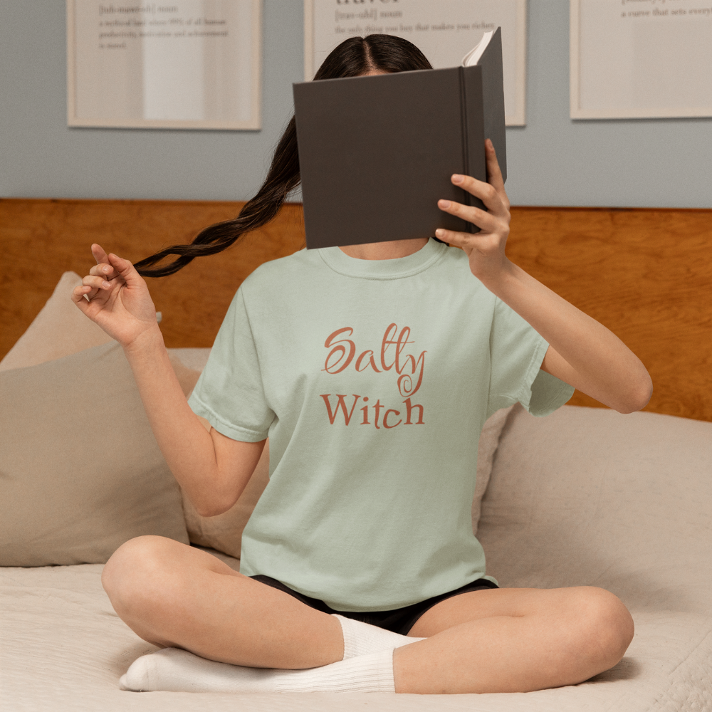 Person sitting on a couch wearing a 'Salty Witch' t-shirt, holding a book over their face.