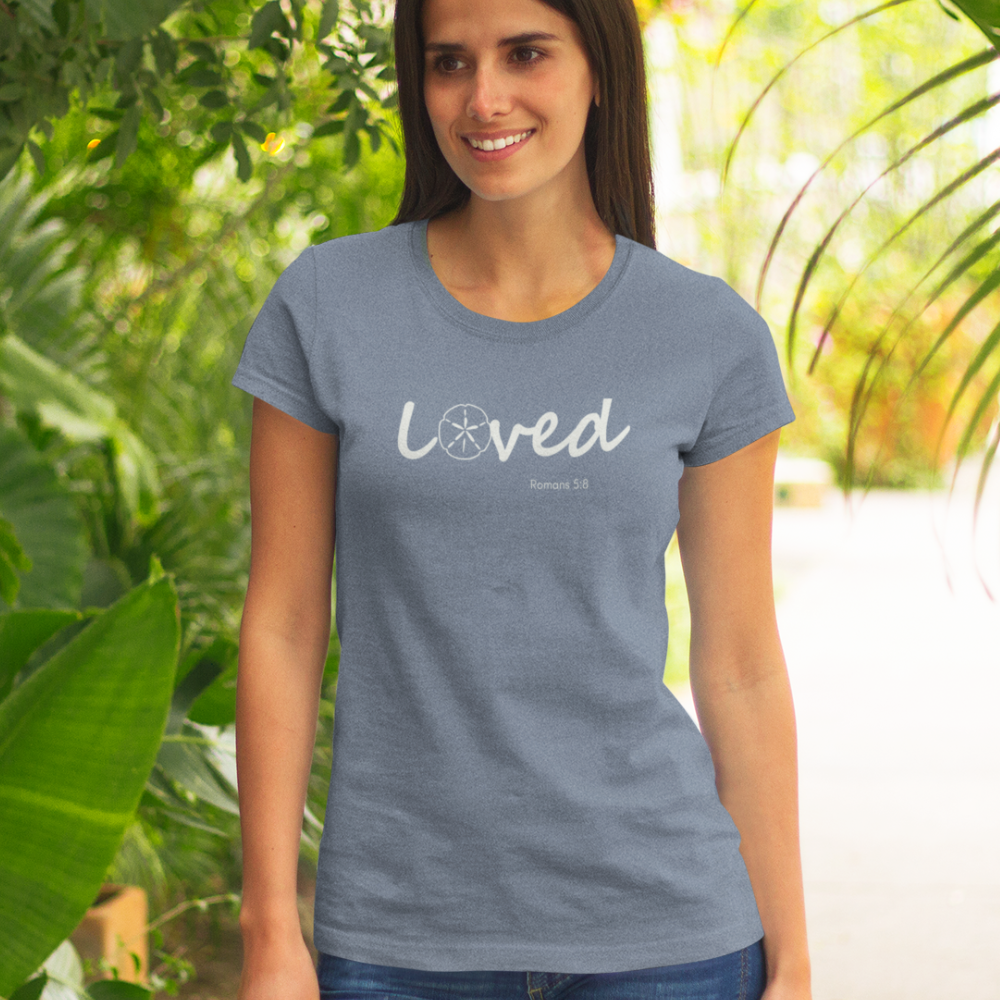 Woman wearing a blue t-shirt with 'loved' printed on it, standing outdoors with greenery.