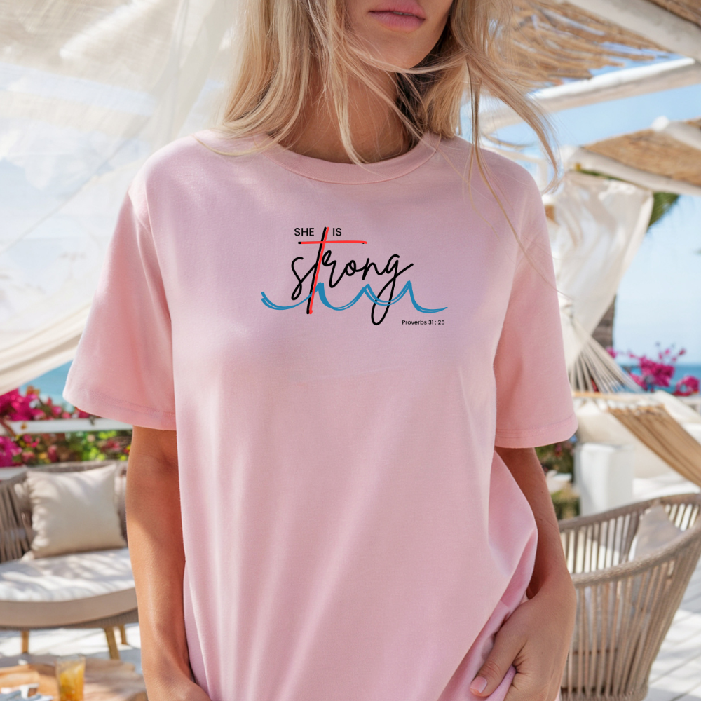 Person wearing a pink t-shirt with 'She is Strong' text at a beachside location.