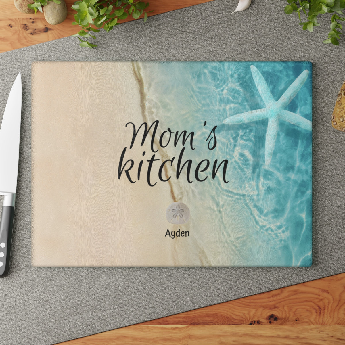 A personalized tempered glass cutting board with a beach-themed design, featuring the text 'Mom's Kitchen' and names of children displayed on sand dollars. The board is placed on a kitchen counter.