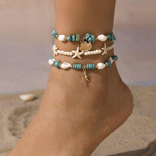 Ankle with multiple beaded bracelets featuring starfish and shell charms on a sandy background.