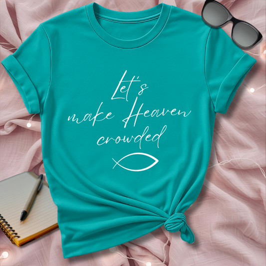 Turquoise t-shirt with 'Let's make Heaven crowded' text on a pink surface with sunglasses and notebook.
