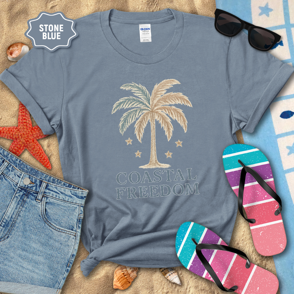 Stone blue t-shirt with palm tree design and 'Coastal Freedom' text, surrounded by beach accessories on sand.