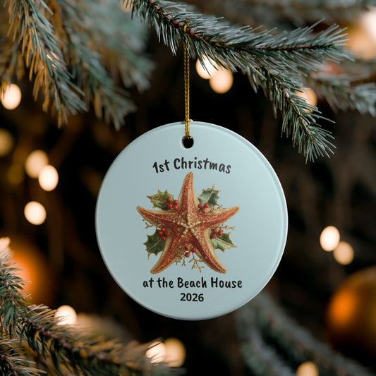 A personalized round Christmas ornament hanging from a pine tree branch. The ornament is light blue with a printed starfish, holly, and the text '1st Christmas at the Beach House 2026'.