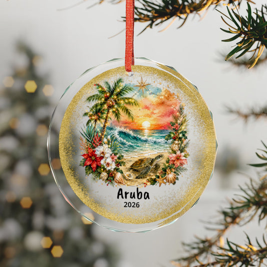 A round clear glass Christmas ornament hanging from a red ribbon, featuring a tropical sunset scene with palm trees, flowers, waves, and a turtle, with the text 'Aruba 2026' printed near the bottom. The ornament is photographed against a softly blurred holiday tree background with pine branches in the foreground.
