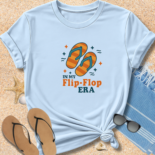 Light blue t-shirt with flip-flop design and text on sand with beach items.