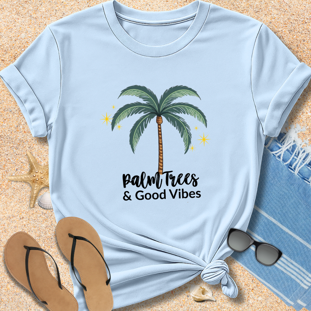 Light blue t-shirt with palm tree design and 'Palm Trees & Good Vibes' text on sand with beach accessories.