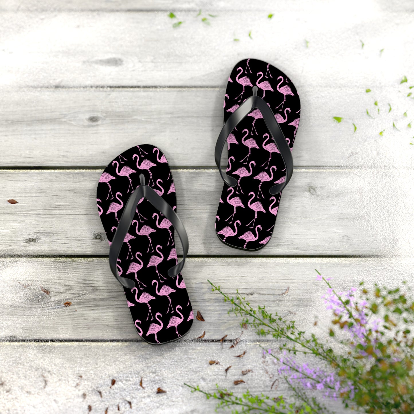 Black flip-flops with pink flamingo pattern on a wooden surface with flowers.