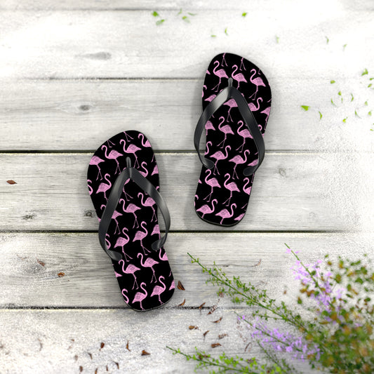 Black flip-flops with pink flamingo pattern on a wooden surface with flowers.