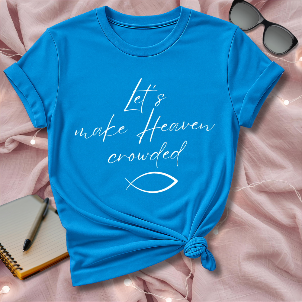 Blue t-shirt with 'Let's make Heaven crowded' text on a pink surface with sunglasses and notebook.