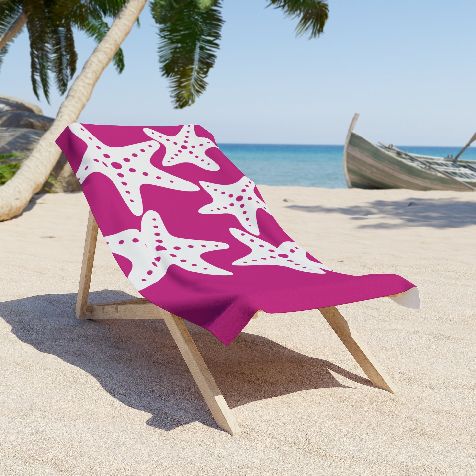 Pink beach towel with white starfish pattern on a wooden chair on a sandy beach.