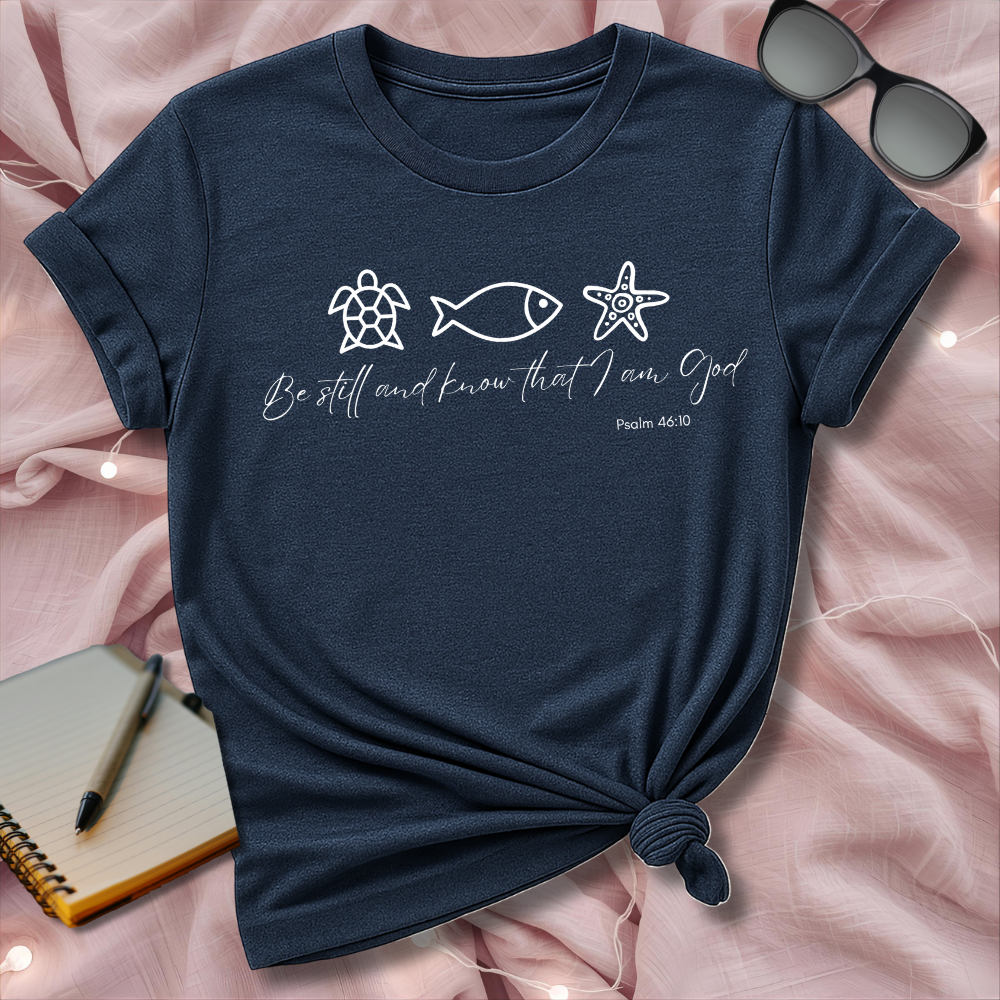 Navy blue t-shirt with sea-themed design and text on a pink surface with sunglasses and notebook.