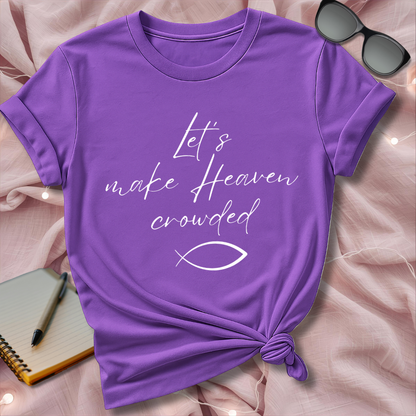Purple t-shirt with 'Let's make Heaven crowded' text and fish symbol on a pink surface.