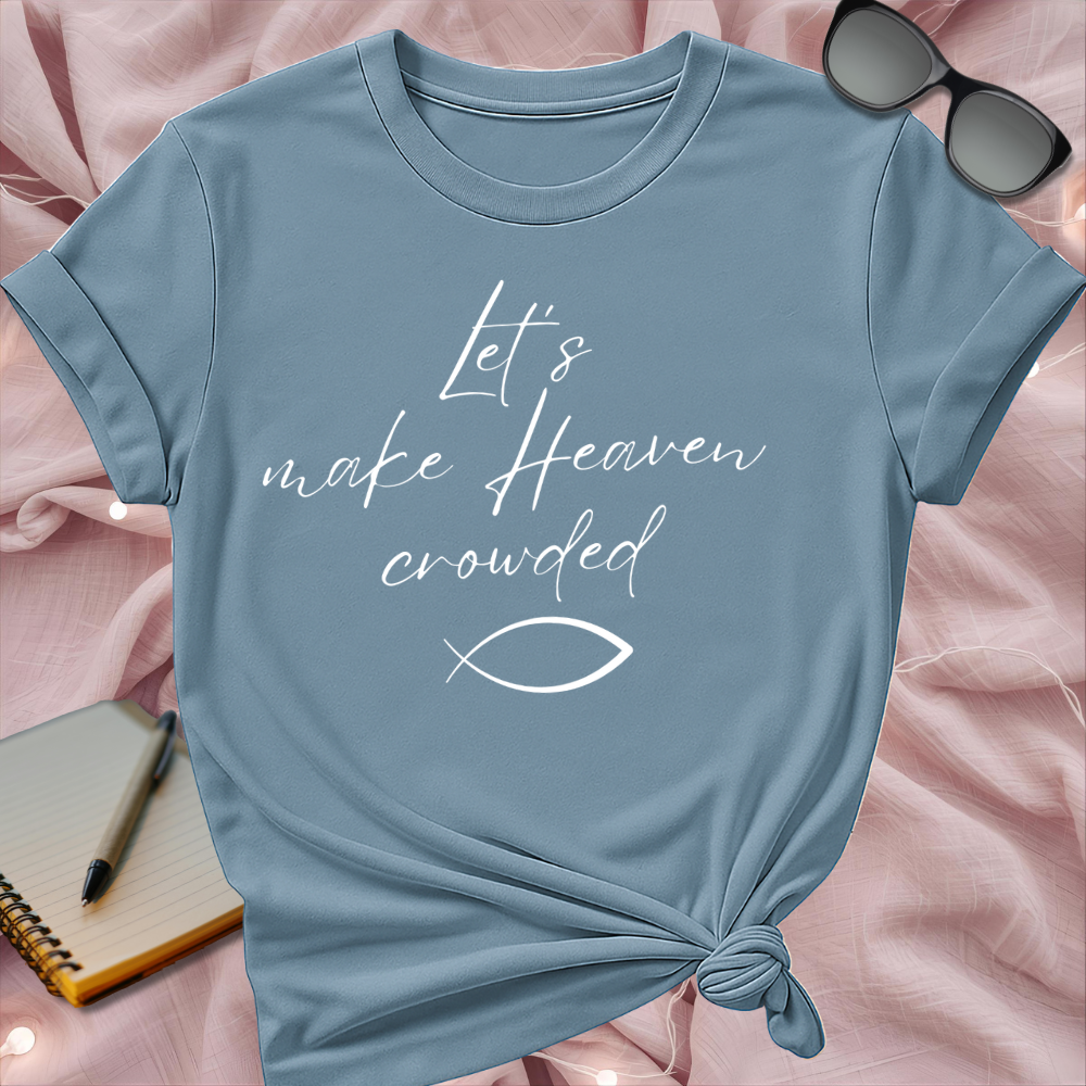 Blue t-shirt with 'Let's make Heaven crowded' text on a pink surface with sunglasses and notebook.
