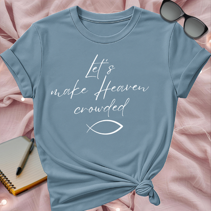 Blue t-shirt with 'Let's make Heaven crowded' text on a pink surface with sunglasses and notebook.