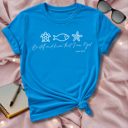 Blue t-shirt with religious text and symbols on a pink surface with sunglasses and notebook.