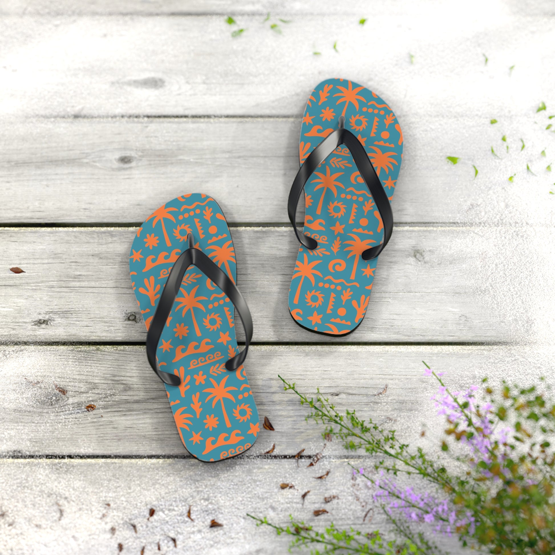 Pair of blue flip-flops with orange patterns on a wooden surface with flowers.