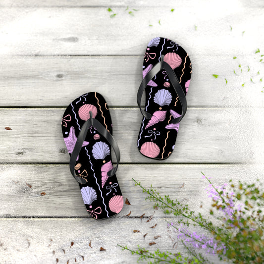 Black flip-flops with pink and purple shell pattern on a wooden surface with flowers.
