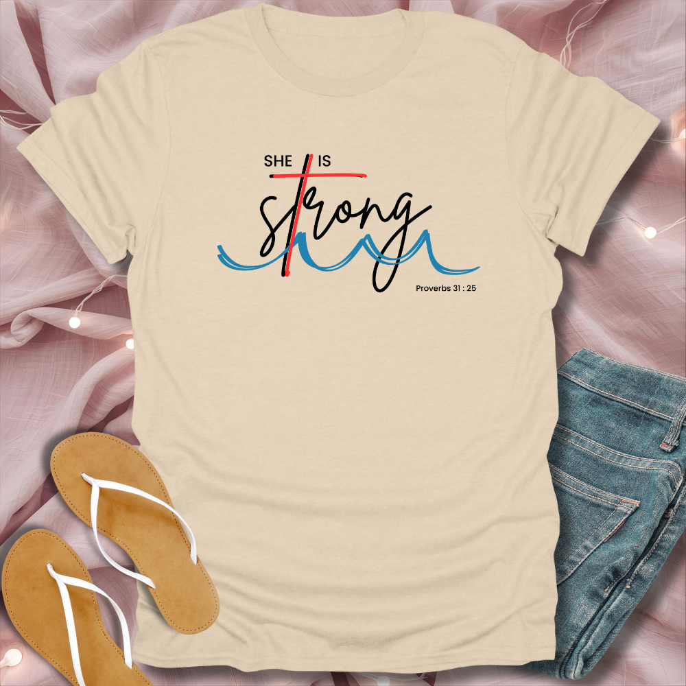 Beige t-shirt with 'She is Strong' text on a pink surface with jeans and sandals.
