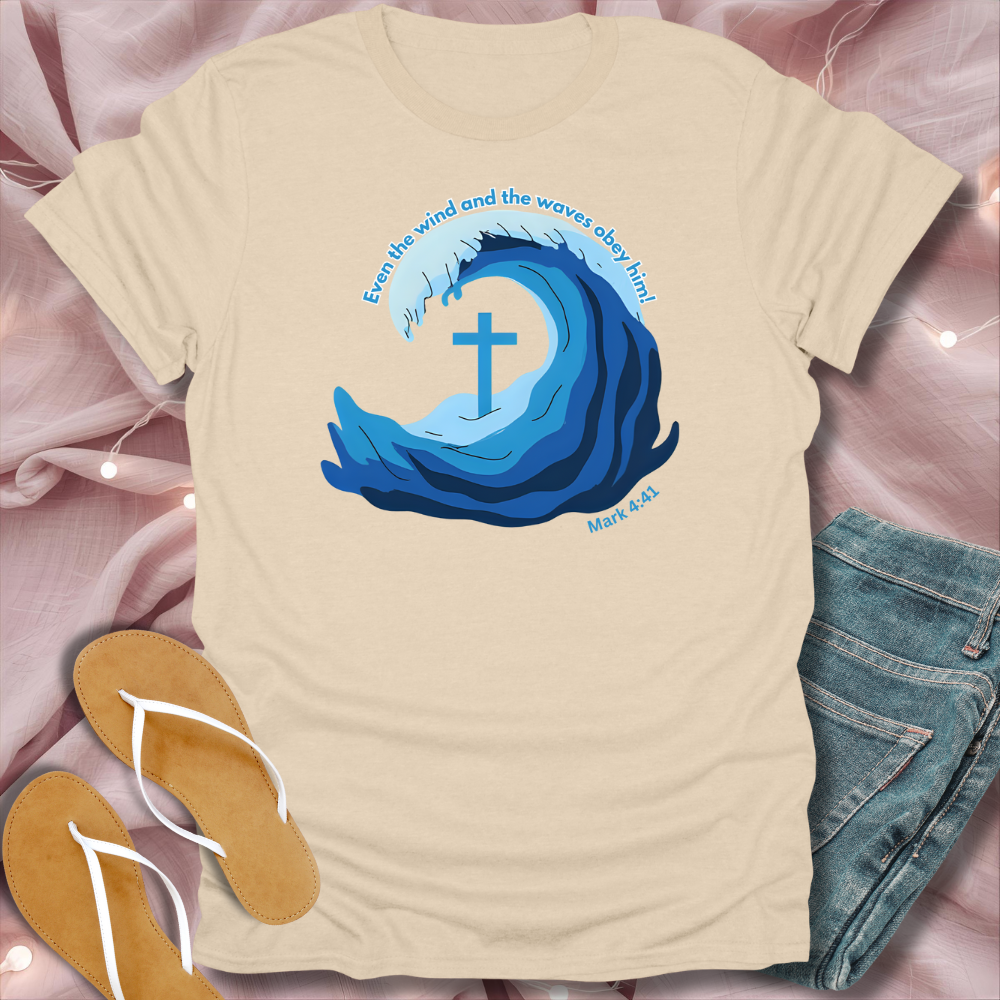 Beige t-shirt with blue wave design and cross, placed on a pink surface with jeans and sandals.