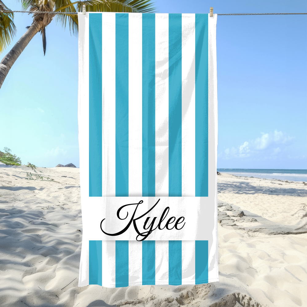 Beach towel with blue and white stripes and 'Kylee' branding on a sandy beach with palm trees.