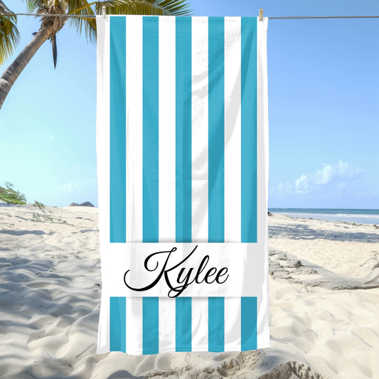 Beach towel with blue and white stripes and 'Kylee' branding on a sandy beach with palm trees.