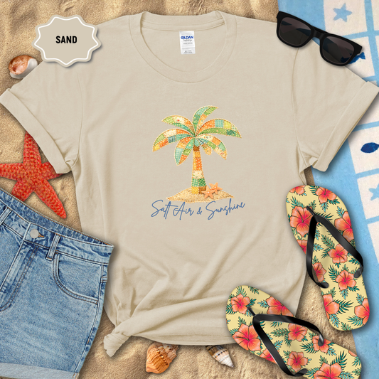 Beige t-shirt with palm tree design and text on sand, surrounded by beach accessories.