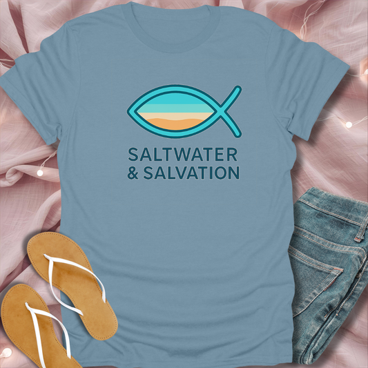 Blue t-shirt with fish symbol and 'Saltwater & Salvation' text on a pink surface with flip-flops and jeans.