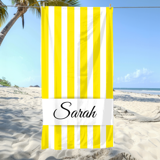 Beach towel with yellow and white stripes and 'Sarah' printed on it, placed on a sandy beach with palm trees.
