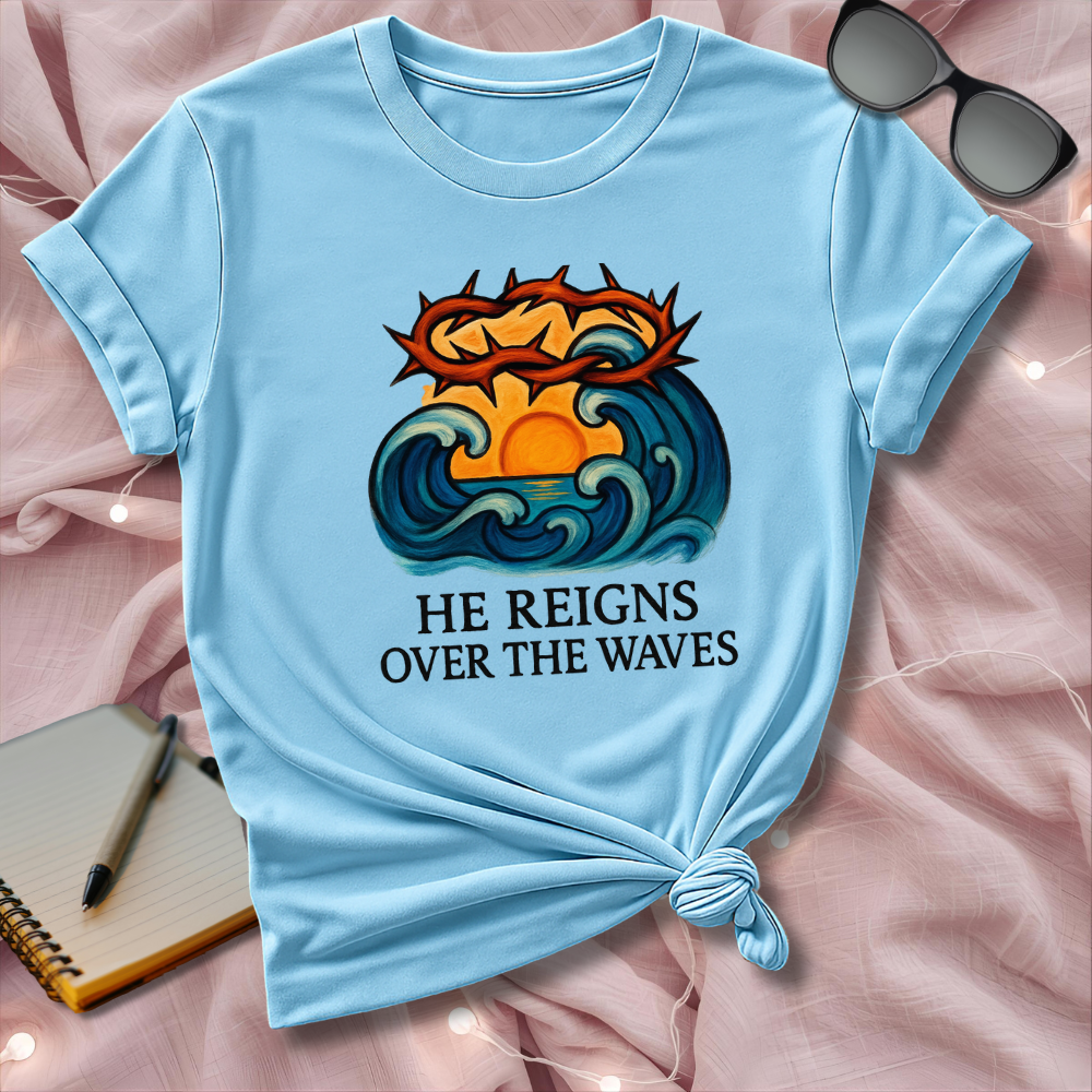 Light blue t-shirt with a graphic design and text 'He Reigns Over the Waves' on a pink surface.