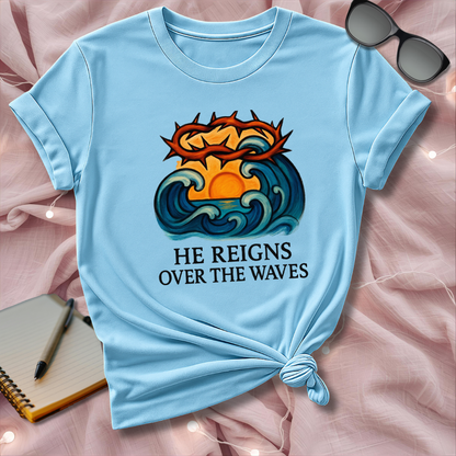 Light blue t-shirt with a graphic design and text 'He Reigns Over the Waves' on a pink surface.