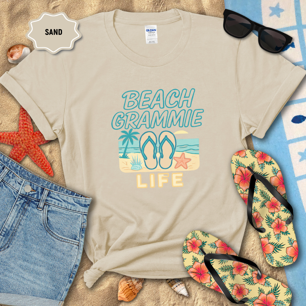 Beige t-shirt with 'Beach Grammie Life' design on sand with flip-flops and sunglasses.
