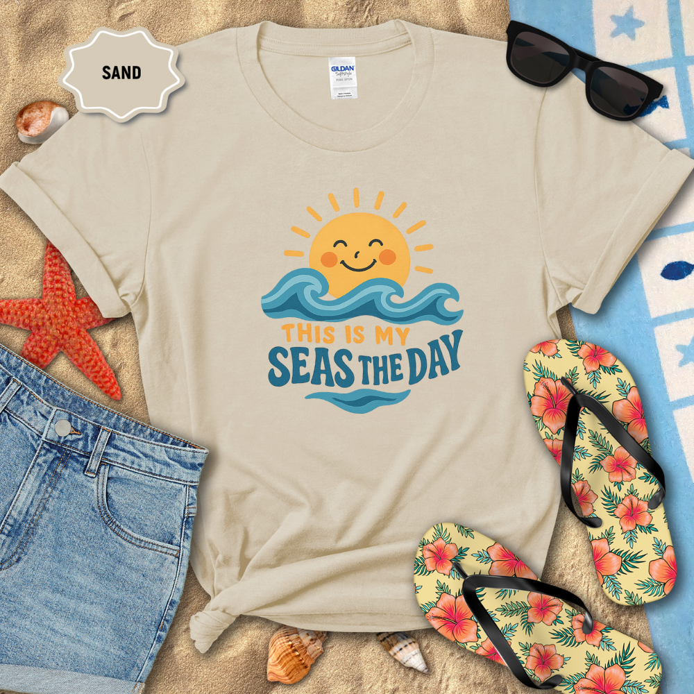 Beige t-shirt with 'This is my seas the day' design on sand with beach accessories.