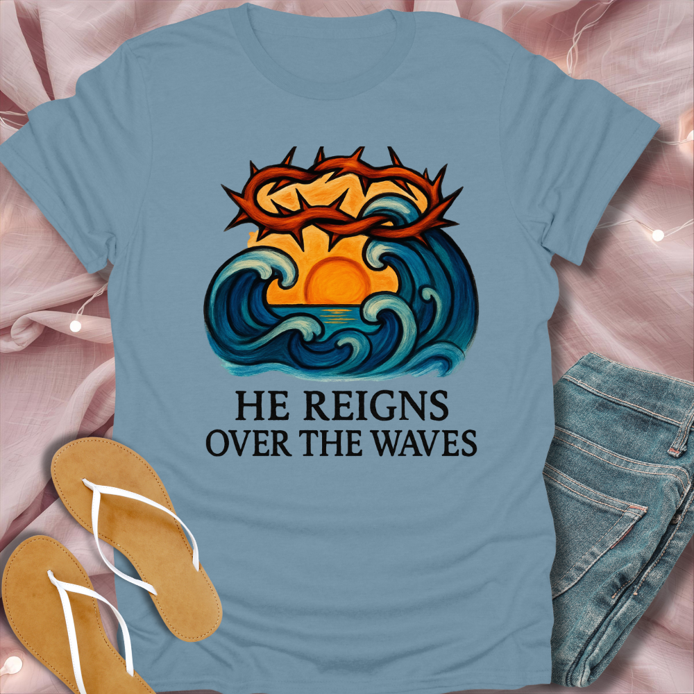 Blue t-shirt with a graphic design and text 'He Reigns Over the Waves' on a pink fabric background.