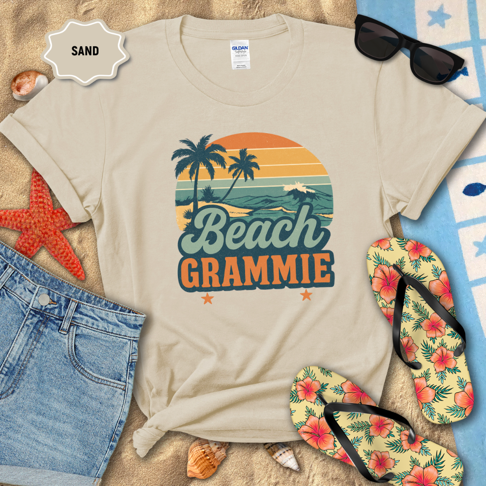 Beige t-shirt with 'Beach Grammie' design on sand with sunglasses, flip-flops, and beach accessories.