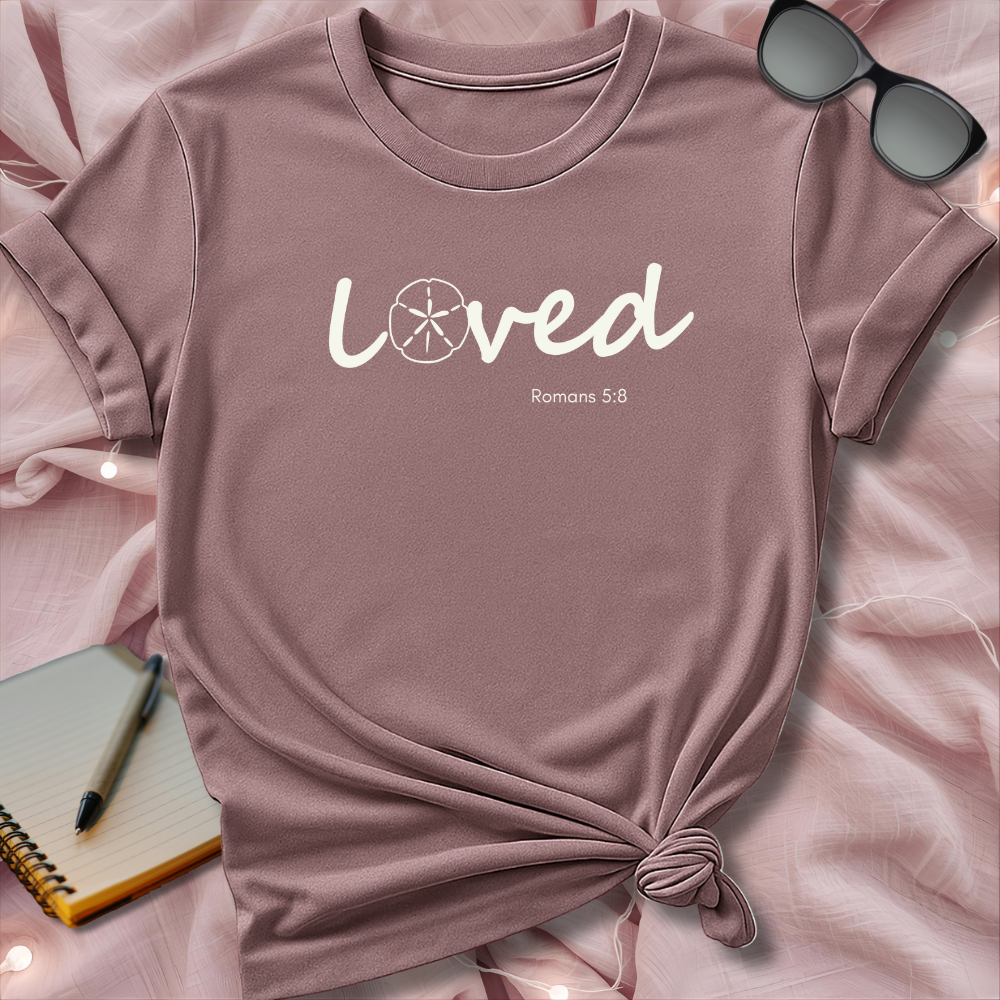 Pink t-shirt with 'Loved Romans 5:8' text on a pink surface with sunglasses and notebook.