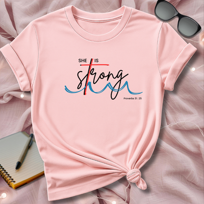 Pink t-shirt with 'She is Strong' text on a pink surface with sunglasses and notebook.