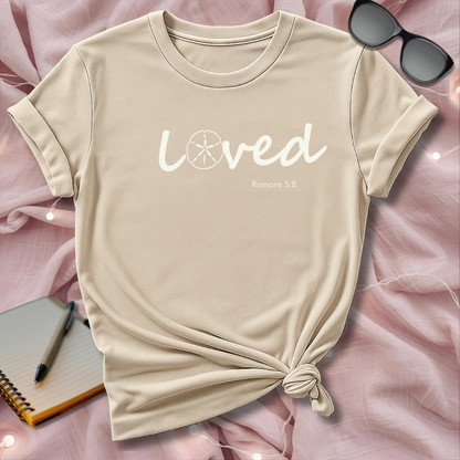 Beige t-shirt with 'loved' text on a pink surface with sunglasses and notebook.