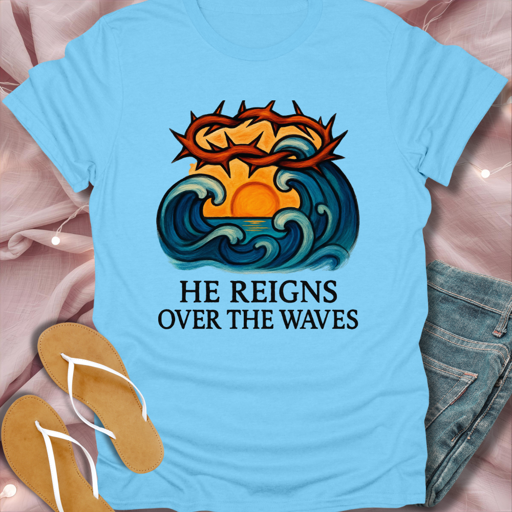 Light blue t-shirt with a graphic design and text 'He Reigns Over the Waves' on a pink fabric background.