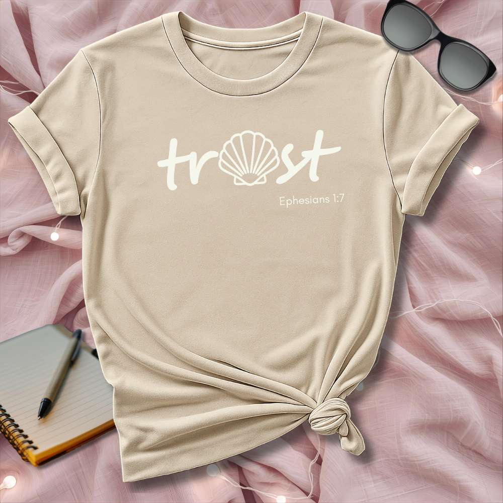 Beige t-shirt with 'trust' text and shell graphic on a pink surface with sunglasses and notebook.