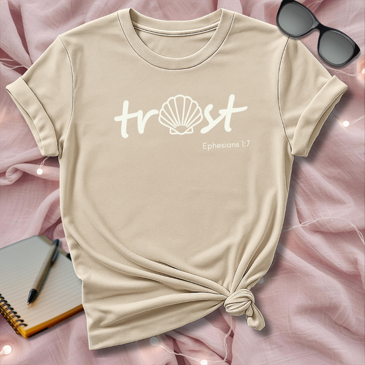 Beige t-shirt with 'trust' text and shell graphic on a pink surface with sunglasses and notebook.
