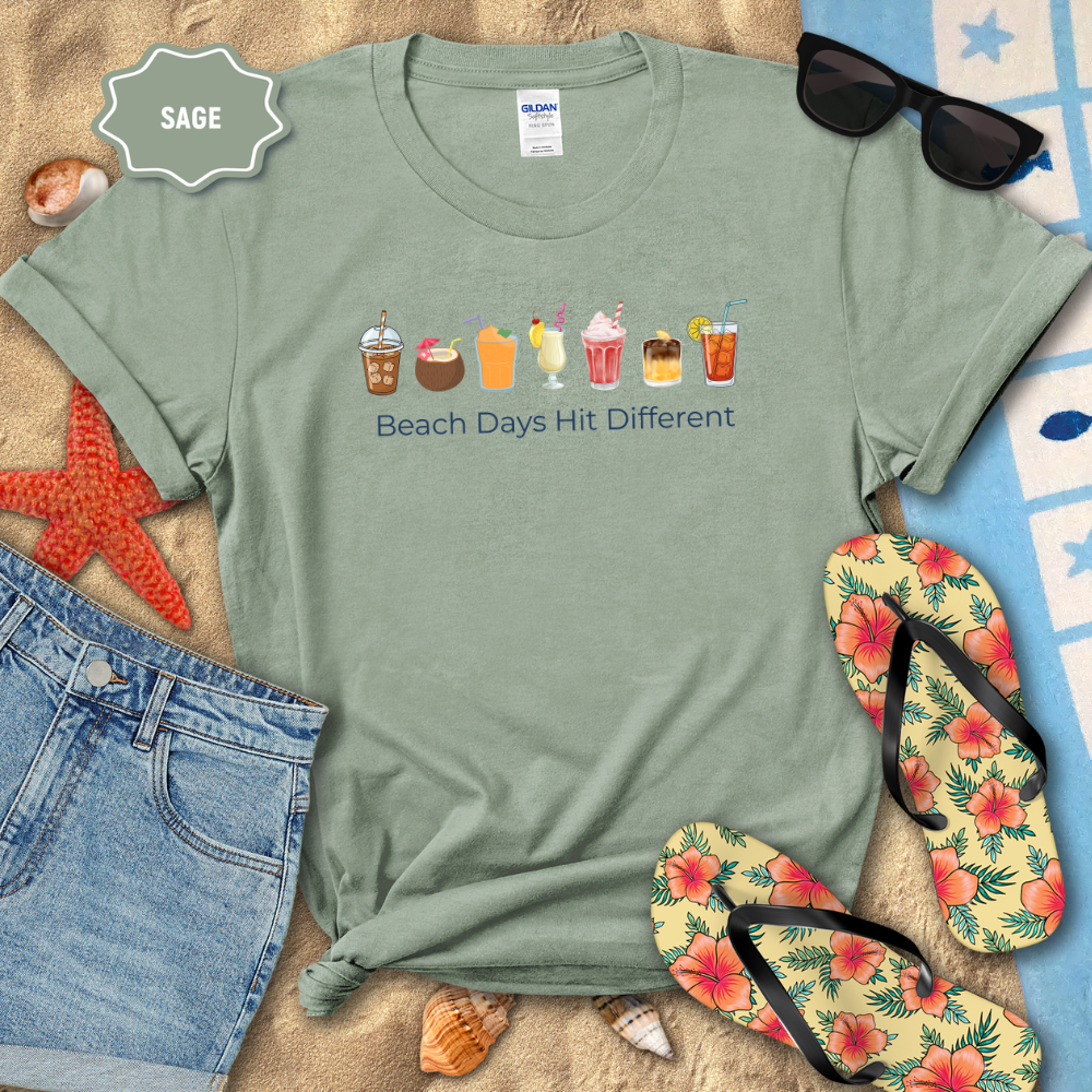 Sage green t-shirt with beach-themed graphics and text, surrounded by beach accessories on sand.