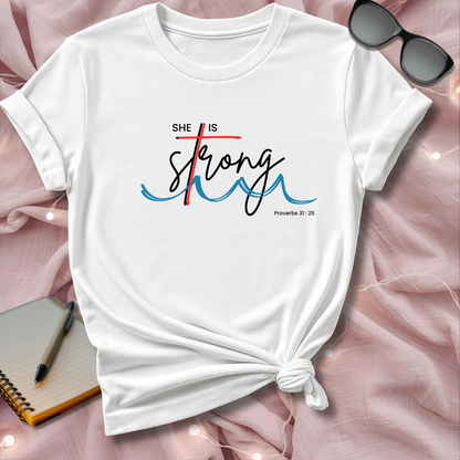 White t-shirt with 'She is Strong' text on a pink surface with sunglasses and notebook.