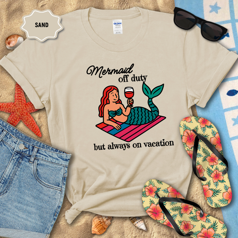 Beige t-shirt with mermaid graphic and text on a sandy background with beach accessories.