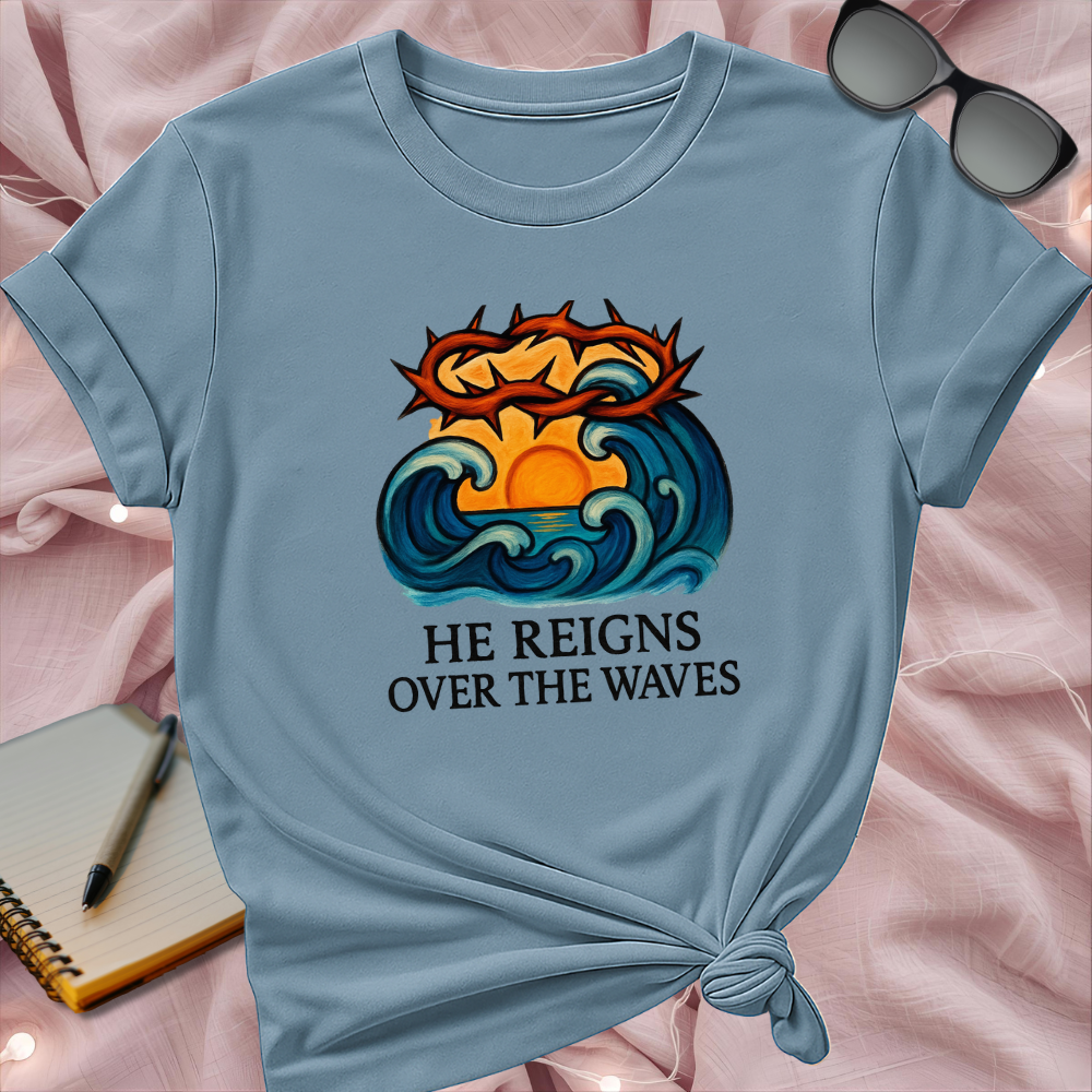 Light blue t-shirt with a graphic design and text 'He Reigns Over The Waves' on a pink surface.