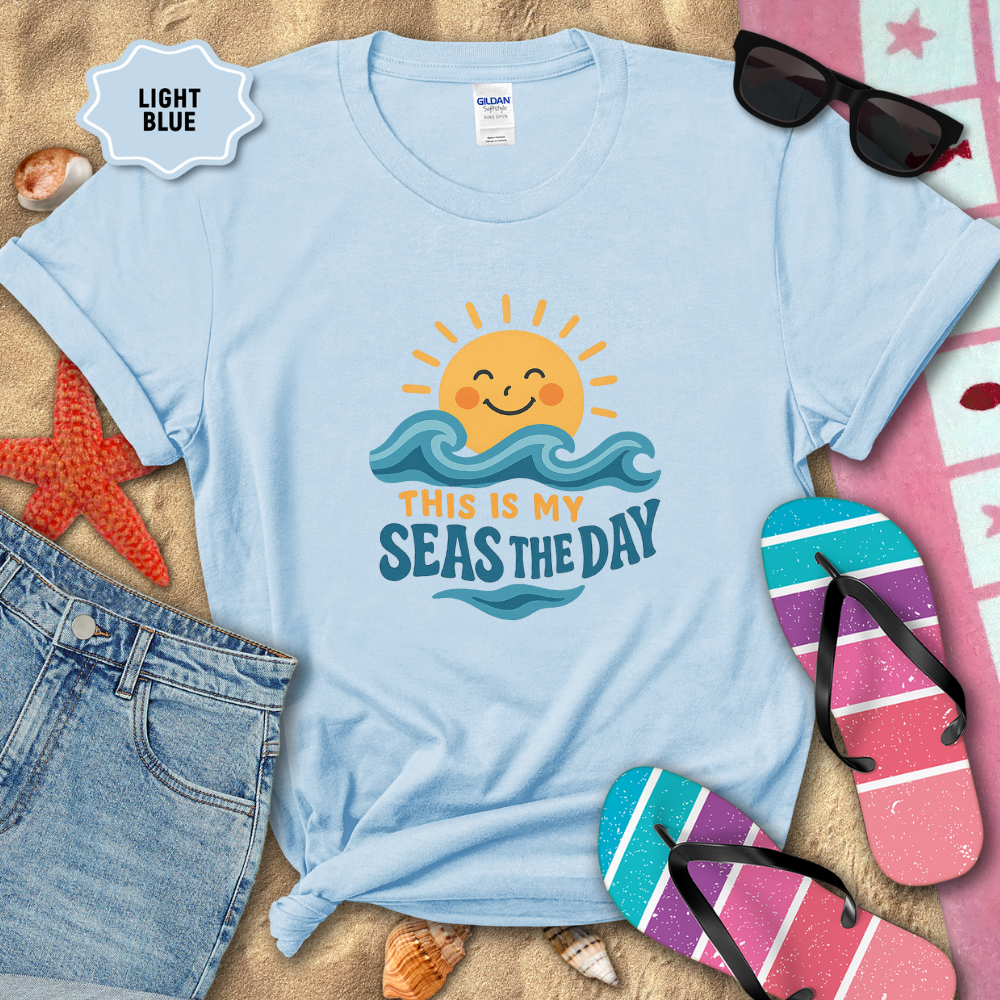 Light blue t-shirt with beach-themed design on sand with sunglasses, flip-flops, and jeans.